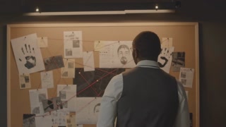 Medium back view of African American detective standing by evidence cork board with criminal case info filled with suspects identikits and handprints, clippings from newspaper and crime scene photos