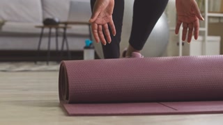 Low section of unrecognizable Black woman in activewear rolling up mat after finishing sports training at home
