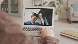 Back view of unrecognizable aged man sitting at desk at home in front of laptop and having video chat with his son wearing graduation gown and hat