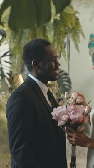 Vertical medium shot of beautiful young newlywed Black couple kissing after wedding ceremony, standing in beautiful room filled with tropic plants