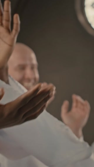 Vertical medium shot of multiethnic male church choir in white gowns clapping hands and singing song while making performance in Catholic Church