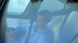 Male businessman reflecting in panoramic roof of modern electric car during browses mobile phone. Young entrepreneur using phone while