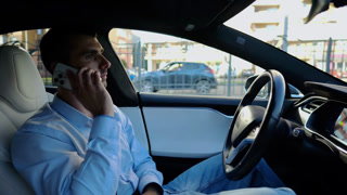 Male entrepreneur speaking by smartphone during riding on electrical auto with autopilot at urban road. Confident businessman talking on phone while riding an autonomous self driving electric car