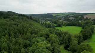 Aerial view of verdant fields and dense woodlands meeting a quaint village under a cloudy sky, creating a serene landscape, Erezee, Region wallonne, Belgium.