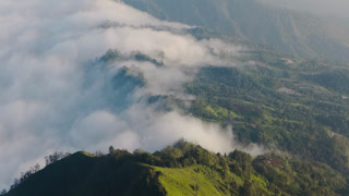 Aerial view of the vibrant green mountain range, partially concealed by a blanket of soft, white clouds, creating a serene and ethereal landscape, Bali, Indonesia.