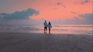 Cinematic shot of loving couple back view walking hand in hand toward sea at sunset. Romantic date on the beach as man gently kisses woman