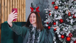 Cheerful young woman Wearing festive antlers talking via video call on her smartphone, sitting near a Christmas tree with bright ornaments