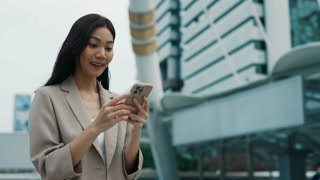 Portrait of Thai businesswoman happy of unexpected financial bonus against office building. Asian lady checks mobile banking app outdoors
