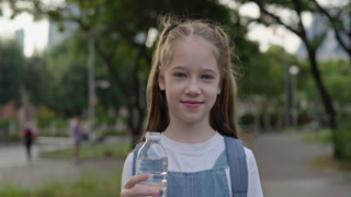 Portrait of pretty junior schoolgirl holds water bottle in green city park. Little girl elementary student with drink stands on town street