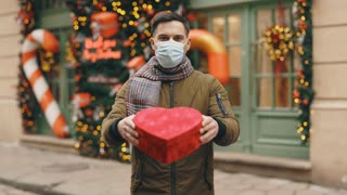 Portrait of the caucasian man in medical mask standing in the decorated street holding the red heart shape gift for the valentines day. Male