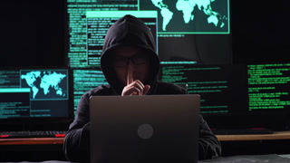 Asian Male Hacker In A Hood Showing Shh Gesture With Index Finger Near Mouth While Works On A Laptop With Maps And Data On Display Screens Of Multiple Monitors In A Dark Office Room
