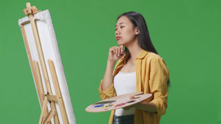 Asian female artist holding her chin and thinking over picture concept while painting on canvas by oil paints and brush in the green screen studio