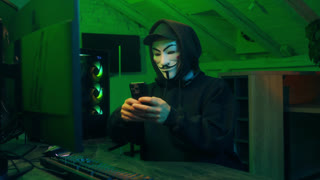 Lviv, Ukraine - June 03, 2024: Anonymous hacker smartphone monitor computer technology cyber crime thief masked man danger pointing finger