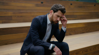 Sad Caucasian businessman in suit thinking about problem with stressed expression while sitting in urban park