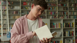 Caucasian teen man guy student choosing between book and mobile phone in library male teenage boy schoolboy confused puzzled pupil choose
