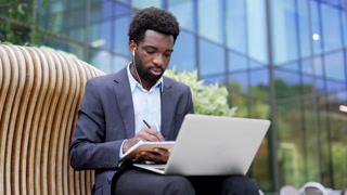 African american businessman watching video call online conference notes in notebook while sitting on a bench on street near office building. Worker in headphones on remote business meeting, training