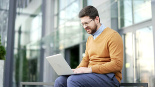 Frustrated stressed businessman reading bad news on laptop computer while sitting on a bench on street near an office building. Shocked worried entrepreneur has a stress looking at negative message