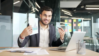 Confident manager presenting chart data at workspace in office. Businessman in a formal suit discussing growth statistics by video call using laptop. Business meeting, analysis, and teamwork concepts