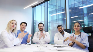 Group of doctors applauding look at camera sitting in a hospital office boardroom celebrate success. Happy medical professional physicians in white coats diverse team colleagues rejoice achievement