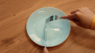 Hands hold a knife and fork positioned on a light blue plate at a wooden table. The scene captures the moment before a meal, emphasizing