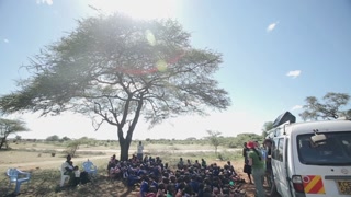 KENYA, KISUMU - MAY 20, 2017: African children sitting on the ground near tree and listen to Caucasian men and women.