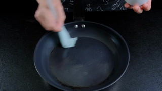 Hands scrub a dark gray frying pan with a light blue brush in soapy water on a black stovetop, indoors in a kitchen during daytime, Cooking location.