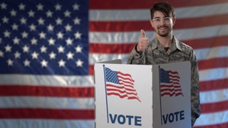 Conceptual portrait US flag background with smiling teen man giving thumbs up, in voting booth at ballot box, careful decision for president in US election.