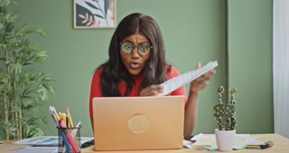 Angry black businesswoman making video call. Mad African American female boss in glasses showing papers and scolding employee while sitting at desk and making video call via laptop in workplace