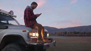 Male traveler is making route, while is sitting on a car hood against mountains view. Caucasian man, traveler and adventurer with a beard