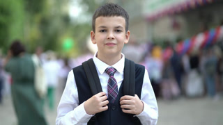 Portrait of smiling schoolboy in uniform and backpack standing near school with classmates and parents blurred in the background.