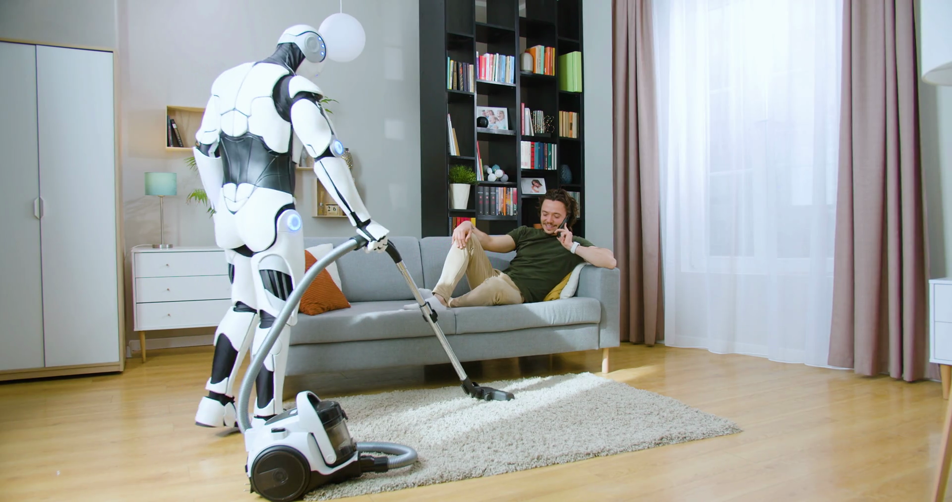 Humanoid robot vacuuming carpet while man in casual wear lying on grey