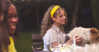 Young Caucasian girl sitting at table and holding cute dog at party in garden at night. Joyful multi-ethnic friends having fun
