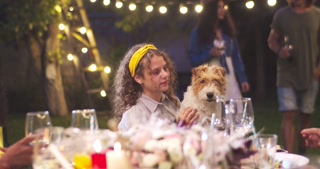 Young cheerful Caucasian girl sitting at table and holding cute dog. Multi-ethnic friends having fun at party in garden at night