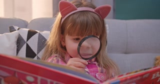 Close-up of adorable child in cute ears reading book in living room. Kid explores textbook with magnifying glass at home.