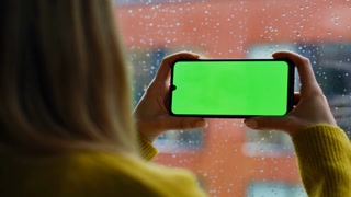 Hands holding horizontal greenscreen smartphone at home closeup. Unrecognizable woman watching chroma key mobile phone standing by rainy