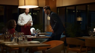 Restaurant waiter serving couple on fancy evening dinner date. Multiethnic lovers walking luxury hotel cafe place. Two people sitting ordering choosing food menu at bar table. Romantic time concept.