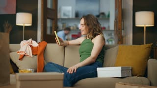 Satisfied shopaholic holding telephone at messy house. Happy girl looking thinking of new purchases. Smiling woman creating shopping list at mobile phone app in apartment. Carefree lifestyle concept