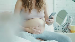 Close up pregnant belly woman sitting with smartphone in bed . Closeup pregnant woman taking selfie photo of tummy at home. Belly mother showing thumb up gesture for photo indoors.