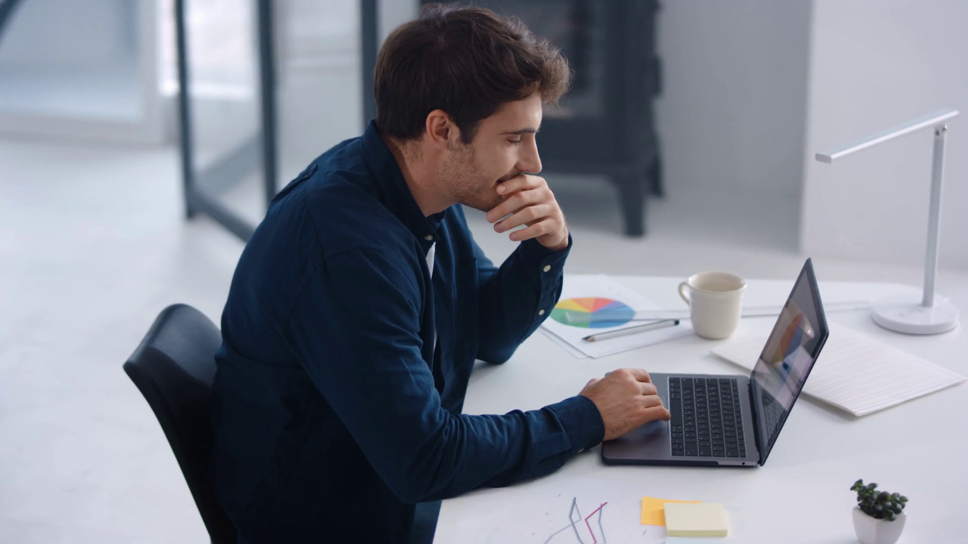  A man in business-casual clothing sits at his desk looking at his laptop screen while holding his chin in thought with a cup of coffee and papers on the desk.