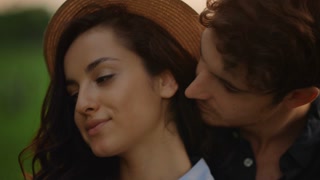 Couple in love expressing tenderness on meadow at sunset. Portrait of charming man hugging woman behind outdoors. Closeup romantic girl and guy spending time together at romantic date in summer park.