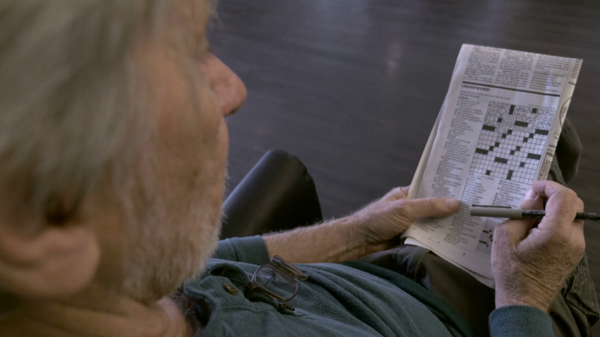 An Elderly Senior Sits In A Chair With A Crossword Puzzle In A 