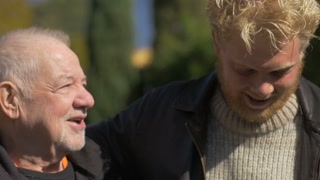 A millennial blond man and an elderly senior talking and embracing outside with their arms on each other's shoulders in the fall or early spring. The older man looks like he is giving advice or saying goodbye.
