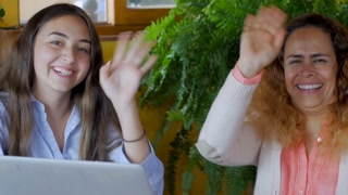 Portrait of a latino mom and teenager daughter waiving and smiling at the camera sitting behind a laptop computer together- slow motion