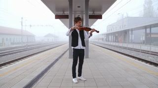 Playing violin on depressed empty train station in slow motion 4K. Wide shot of train station with an elegant teenager in focus playing violin placed in the center of the frame. Rail tracks on the left and right part.