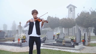 Person play sad violin songs on cemetery in slow motion 4K. Wide shot of a young person with violin in focus stand in the middle of cemetery playing the instrument. Church and graves in the background.