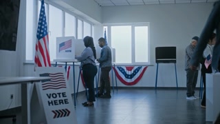 Female polling officer consults male voter, gives ballot for voting. African American man talks to worker at registration table at polling