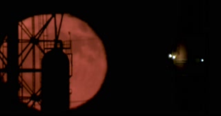 Red full moon rising from behind a large oil refinery