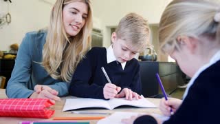 Mother Helping Son And Daughter Wearing School Uniform With Homework At Table In Kitchen