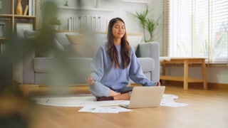 Happy young Asian woman practicing yoga and meditation at home sitting on floor in living room in lotus position and relaxing with closed eyes. Mindful meditation and wellbeing concept