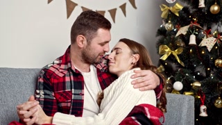 guy tenderly hugs his girlfriend sitting under a warm blanket on the background of the Christmas tree